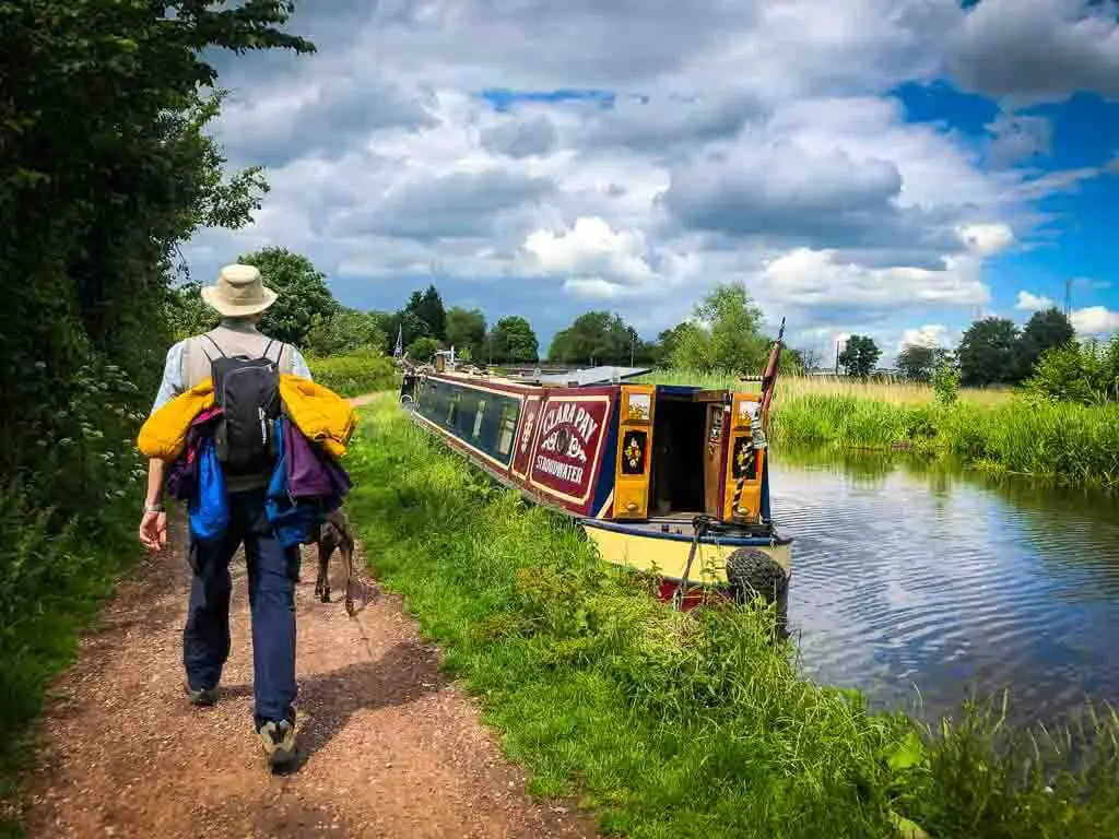 man in hat walking a long a canal path with a brightly painted long boat on the water