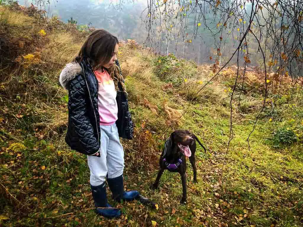 brown dog looking tired next to girl in black coat and fur hood on grassy hill