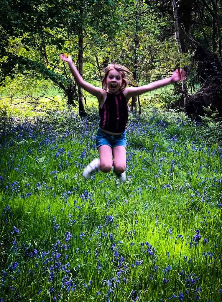 girl jumping in the air with denim shorts on. there are bluebells and grass below her
