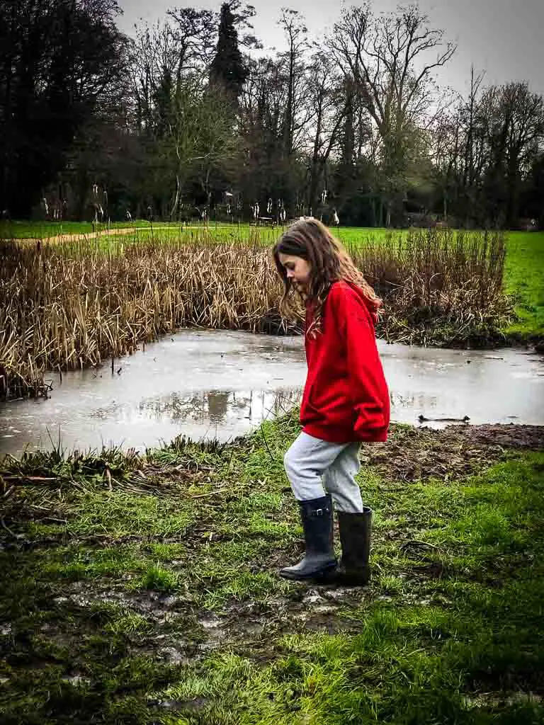 young girls in blue wellies, white jeans, red jumper with long wavy hair stomping in mud infront of a pond