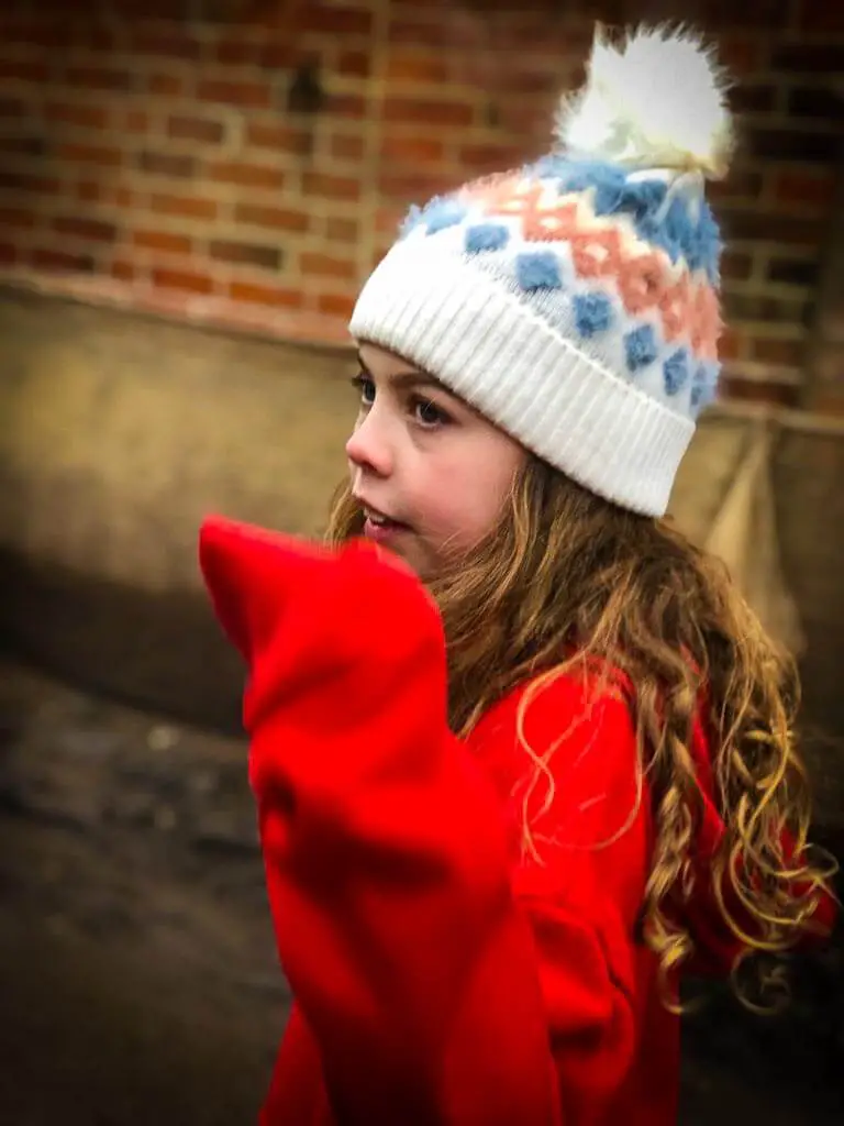 girl with curly blonde hair, red jumper and white wooden hat with pompom on top