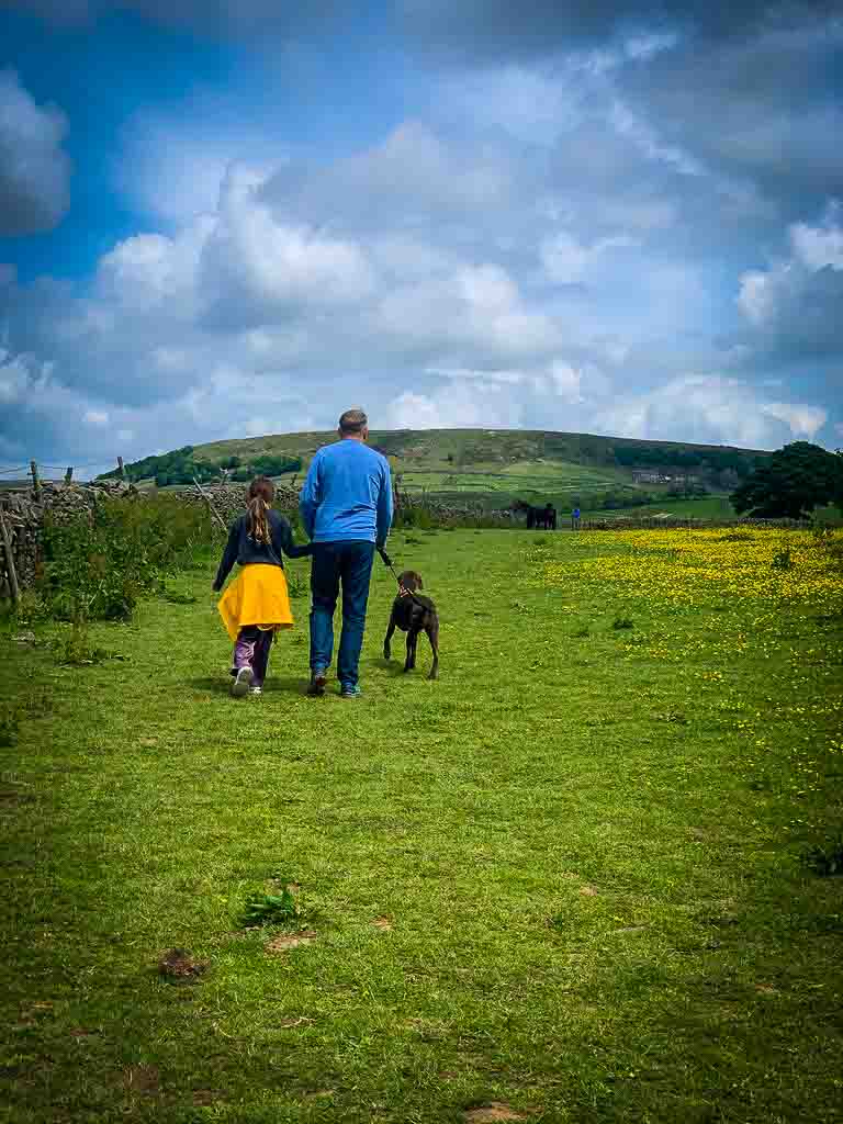 man in blue jumper holding a dog on a lead walking across a green field with a young girl